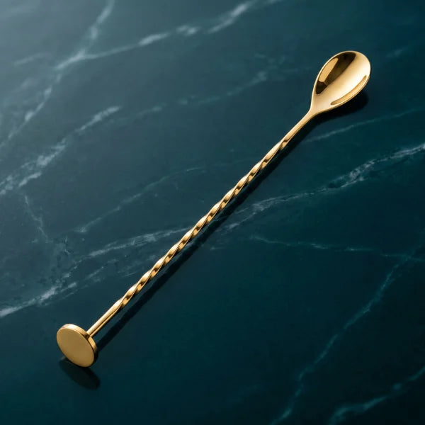 Goldfarbener Barlöffel mit gedrehtem Griff auf dunkelblauer Marmoroberfläche mit heller Maserung, fotografiert im minimalistischen Premium-Stil.