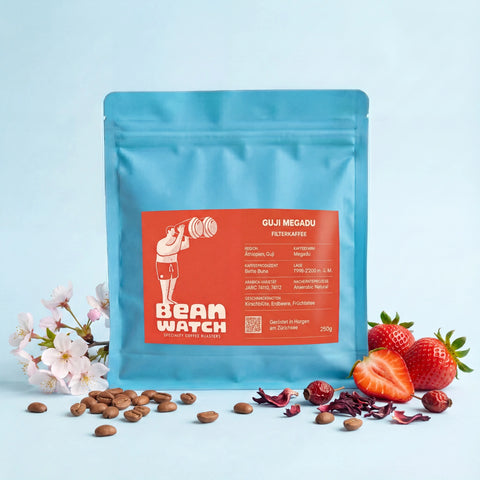 Ein Beutel unserers Guji Megadu Filterkaffee in unserem Beanwatch Studio umgeben von Erdbeeren, Kirschblüten, Früchteteezutaten und vereinzelten, gerösteteten Kaffeebohnen.