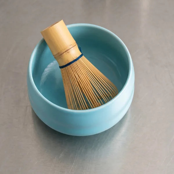 Matcha-Chasen aus Bambus in einer hellblauen Matcha-Schüssel auf einer Edelstahl-Arbeitsfläche, fotografiert im minimalistischen Beanwatch-Stil.